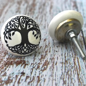 Puede incluir: Pomo de armario de cerámica blanca con un diseño de árbol de la vida negro. El pomo tiene un tornillo plateado.