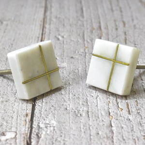 以下が含まれることがあります： 金色の十字架のデザインが施された白い正方形のキャビネットノブが2つ。ノブには、キャビネットドアに取り付けるためのネジが背面にあります。