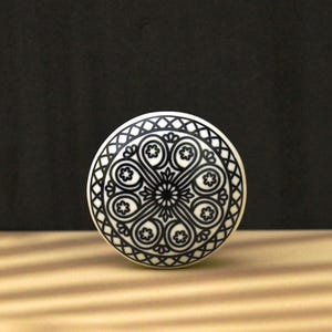Puede incluir: Pomo de cerámica blanco con un diseño geométrico en blanco y negro. El diseño presenta un círculo central con cuatro círculos circundantes, todos con patrones intrincados.