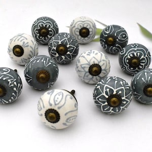 May include: A collection of ceramic cabinet knobs in shades of gray and white, adorned with floral and leaf patterns. Each knob features a brass-colored metal base. The knobs are arranged on a white surface.