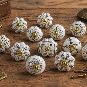 Op de afbeelding: Een verzameling witte keramische kastknoppen met gouden en zwarte bloemmotieven. Verschillende vormen en maten worden getoond op een houten oppervlak, samen met schroeven en een ladeknop.