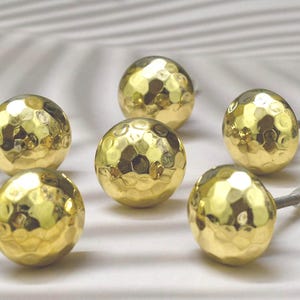Könnte beinhalten: Sechs goldfarbene Schrankknöpfe mit gehämmerter Oberfläche. Die Knöpfe sind rund und haben eine leicht unregelmäßige Oberfläche.