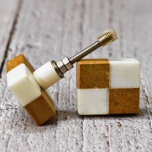 Puede incluir: Dos pomos cuadrados para armario con un patrón de tablero de ajedrez de mármol blanco y marrón. Los pomos tienen un tornillo de metal plateado.