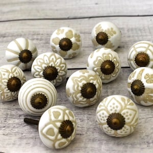 Può includere: Dodici pomelli in ceramica bianca con intricati disegni floreali beige e dettagli in ottone. I pomelli sono disposti su una superficie in legno chiaro.