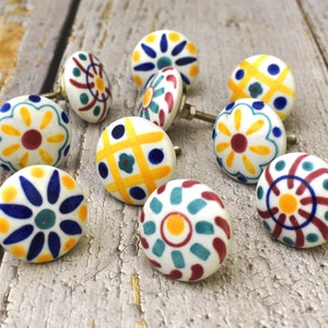 Könnte beinhalten: Sortiment von 10 Keramik-Schrankknöpfen mit bunten floralen und geometrischen Mustern. Die Knöpfe sind weiß mit gelben, blauen, roten und grünen Akzenten.