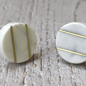 Op de afbeelding: Twee ronde witte marmeren knoppen met gouden afwerking. De knoppen hebben een eenvoudig ontwerp met twee parallelle gouden lijnen.