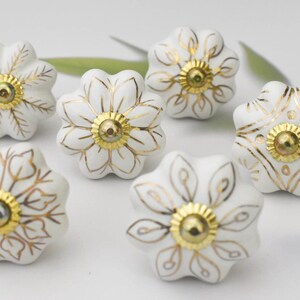May include: Six white ceramic cabinet knobs with gold floral designs. The knobs have a slightly raised, rounded shape.