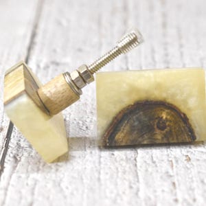 Puede incluir: Dos pomos cuadrados para armario hechos de madera y resina. Los pomos son de color amarillo claro con patrones de veta de madera marrón. Un pomo tiene un tornillo plateado.