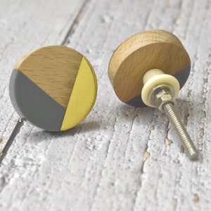 May include: Two round wooden cabinet knobs with a geometric design. One knob is painted gray and gold, the other is painted gray and gold with a silver screw.