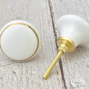 May include: Two white ceramic cabinet knobs with a gold trim. One knob is attached to a gold screw.