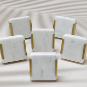 May include: Six white marble cabinet knobs with gold trim. The knobs are square shaped and have a subtle veining pattern.