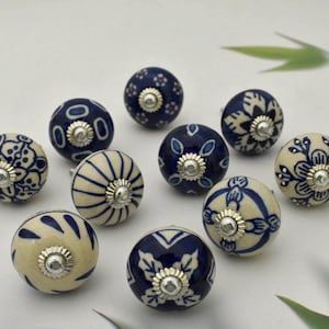 May include: A collection of ten ceramic cabinet knobs. The knobs are round with a cream or navy blue base and feature intricate floral or geometric patterns in contrasting colors. Each knob has a silver-toned metal base.