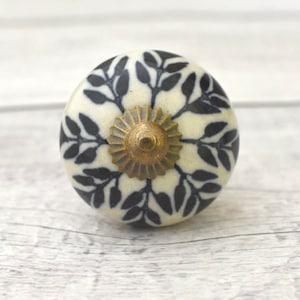 Puede incluir: Pomo de cerámica blanco con un patrón de hojas negras y un centro de metal dorado.