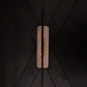 May include: A black incense stick is burning on a rectangular wooden holder. The holder is placed on a dark wood surface with a chevron pattern.