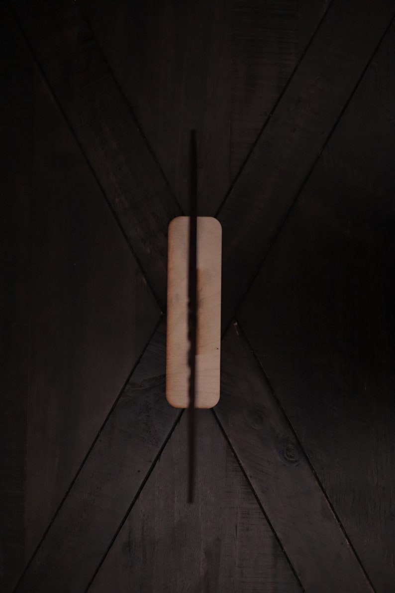 May include: A black incense stick is burning on a rectangular wooden holder. The holder is placed on a dark wood surface with a chevron pattern.
