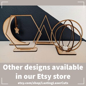 May include: Four wooden geometric shapes, a crescent moon, a hexagon, a triangle, and a sphere, are displayed on a white surface. Each shape has a base and is designed to hold a candle or other decorative item. The text "Other designs available in our Etsy store" and the website address "etsy.com/shop/LastingLaserCuts" are displayed below the shapes.