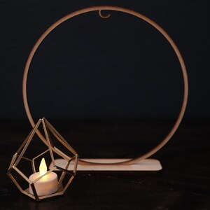 May include: A gold geometric candle holder with a lit candle inside, sitting in front of a wooden circle hoop.