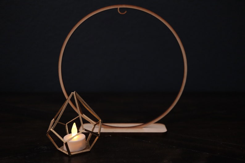 May include: A gold geometric candle holder with a lit candle inside, sitting in front of a wooden circle hoop.