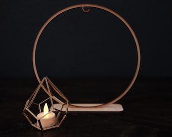 May include: A gold-toned metal geometric candle holder containing a white LED tea light, positioned in front of a large, circular gold-toned metal hoop stand with a flat base, both featuring a metallic finish against a dark background.