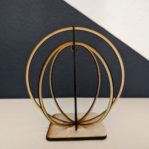 Puede incluir: Una escultura de madera de tres círculos concéntricos, siendo el círculo exterior el más grande y el círculo interior el más pequeño. La escultura está hecha de madera marrón claro y tiene un acabado natural. La escultura está de pie sobre una base cuadrada.