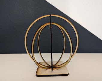 May include: A wooden sculpture of three concentric circles, with the outer circle being the largest and the inner circle being the smallest. The sculpture is made of light brown wood and has a natural finish. The sculpture is standing on a square base.