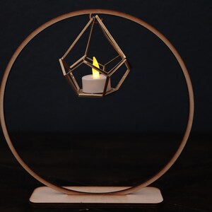 May include: A gold geometric candle holder hangs from a wooden ring stand. The candle holder is suspended in the air and has a small white candle inside.