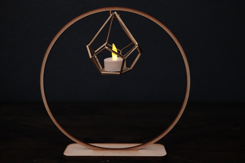 May include: A gold geometric candle holder hangs from a wooden ring stand. The candle holder is suspended in the air and has a small white candle inside.