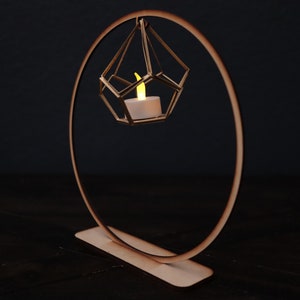 May include: A wooden candle holder with a geometric design. A small, white candle is lit inside the holder, which is suspended from a circular wooden ring.