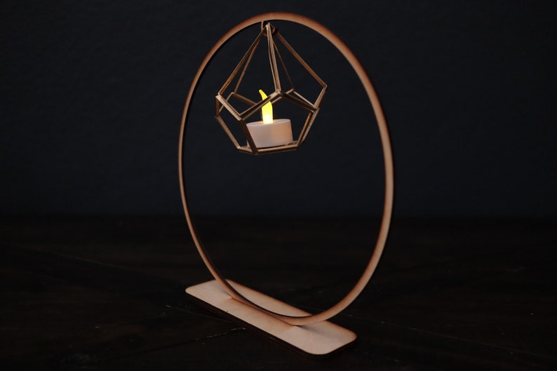 May include: A wooden candle holder with a geometric design. A small, white candle is lit inside the holder, which is suspended from a circular wooden ring.
