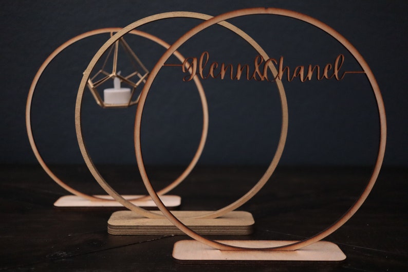 May include: Three wooden circles with a base, the largest circle has the text "Glenn & Chanel" cut out of it. The middle circle has a small gold geometric candle holder hanging from it.