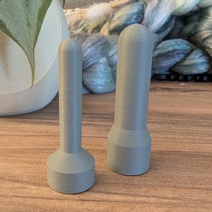 Könnte beinhalten: Zwei graue, 3D-gedruckte zylindrische Werkzeuge mit abgerundeten Oberseiten. Das linke Werkzeug hat einen schmalen Schaft und eine breitere, kreisförmige Basis mit sichtbaren horizontalen Schichtlinien. Das rechte Werkzeug ist dicker.