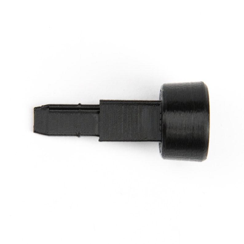 May include: A black plastic push button with a long, thin, rectangular stem. The button is round and has a smooth surface.