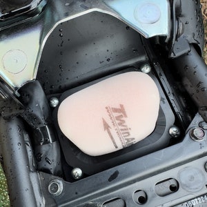 Peut inclure: Gros plan sur un boîtier de filtre à air de moto noir avec un filtre à air beige à l'intérieur. Le filtre à air porte l'inscription "Twin Air".