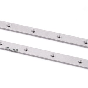 Pode incluir: Duas barras de metal prateadas, retangulares, com v&aacute;rios orif&iacute;cios espa&ccedil;ados uniformemente. As barras t&ecirc;m uma superf&iacute;cie texturizada e est&atilde;o dispostas diagonalmente. O texto "3DLogoGZ" &eacute; vis&iacute;vel numa barra.