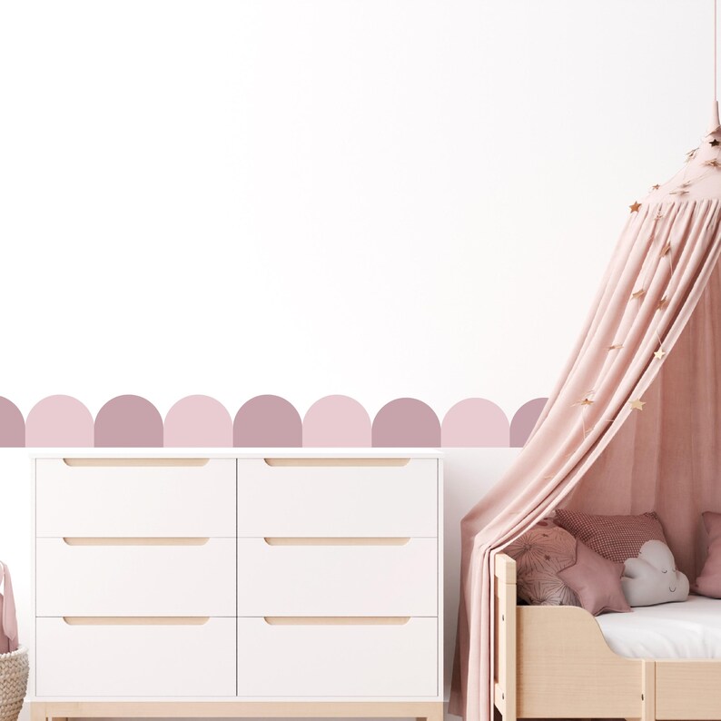 May include: A white chest of drawers with six drawers sits beneath a wall adorned with a row of pink and mauve semi-circles. A canopy bed with a pink canopy and star decorations is on the right.