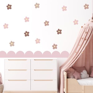May include: A white wall adorned with flower-shaped decals in brown, pink, and beige. A scalloped border in soft pink decorates the wall. A white chest of drawers with six drawers and a wooden bed frame with a pink canopy are also visible.