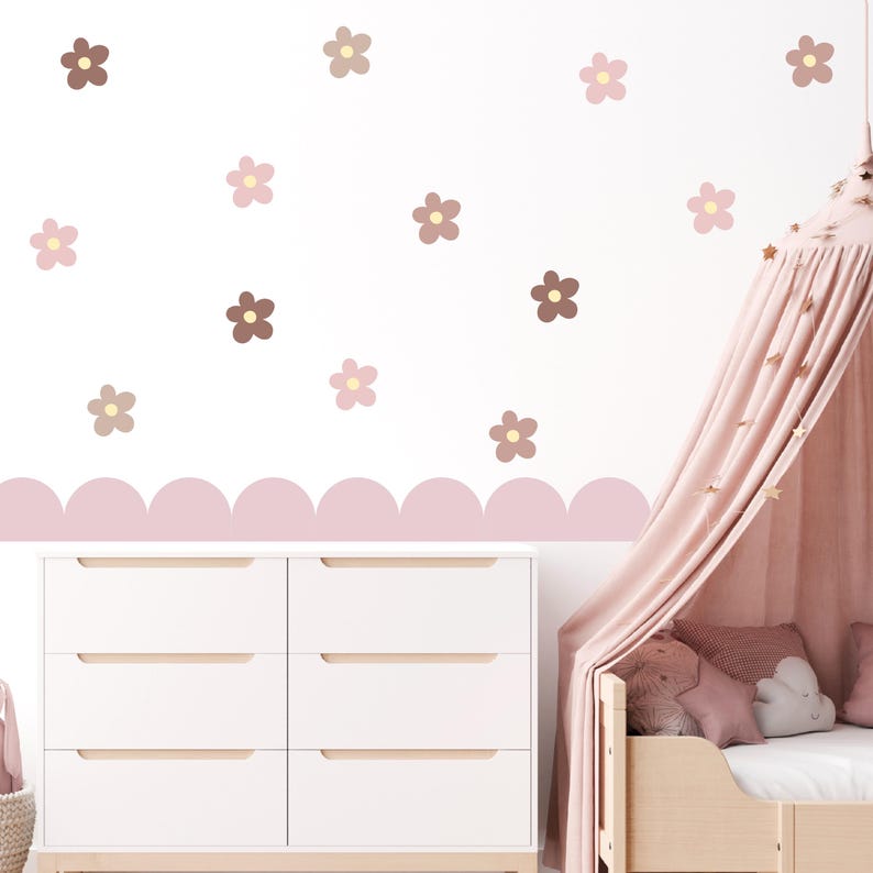 May include: A white wall adorned with flower-shaped decals in brown, pink, and beige. A scalloped border in soft pink decorates the wall. A white chest of drawers with six drawers and a wooden bed frame with a pink canopy are also visible.