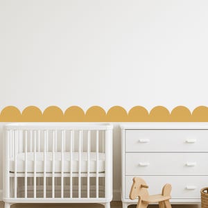 May include: A nursery scene with a white cot, a white chest of drawers, and a mustard-coloured scalloped border on the wall. A wooden rocking horse is in front of the drawers.