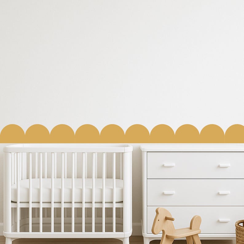 May include: A nursery scene with a white cot, a white chest of drawers, and a mustard-coloured scalloped border on the wall. A wooden rocking horse is in front of the drawers.