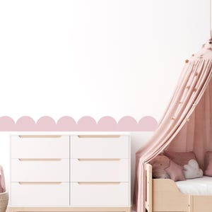 May include: A white six-drawer chest of drawers with light wood handles is positioned below a pink scalloped wall border. A light pink canopy bed with star decorations and pillows is on the right. The room has a soft, pastel colour scheme.