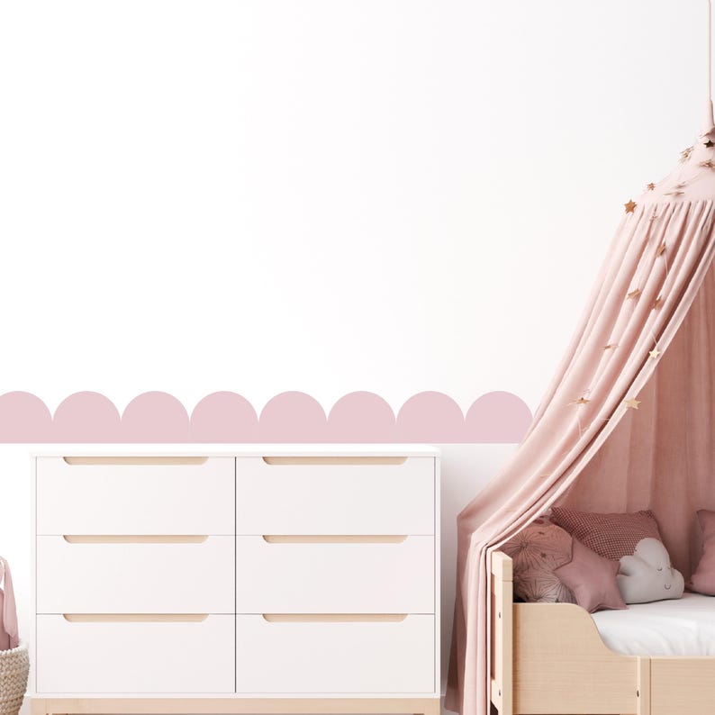 May include: A white six-drawer chest of drawers with light wood handles is positioned below a pink scalloped wall border. A light pink canopy bed with star decorations and pillows is on the right. The room has a soft, pastel colour scheme.