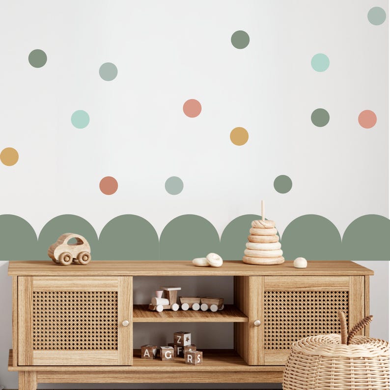 May include: A wooden cabinet with woven doors and a collection of wooden toys. The wall behind the cabinet is white with colourful polka dots and a green scalloped border. Toys include a car, train, stacking rings, and alphabet blocks.