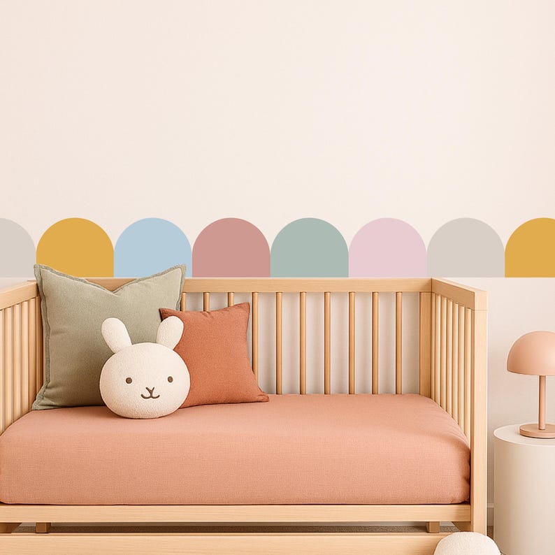 May include: A wooden cot with a peach-coloured fitted sheet, green and rust-coloured cushions, and a bunny-shaped plush toy. The wall above the cot features a row of colourful, semi-circular decals in pastel shades.