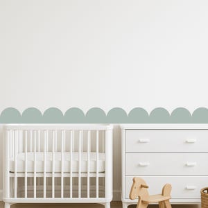 May include: A nursery scene with a white cot, a white chest of drawers, and a wooden rocking horse. A decorative scalloped border in a muted green-blue colour is on the wall above the furniture, creating a soft, inviting atmosphere.