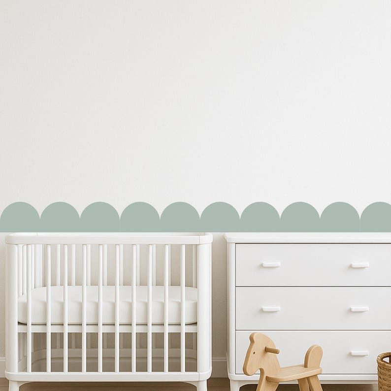 May include: A nursery scene with a white cot, a white chest of drawers, and a wooden rocking horse. A decorative scalloped border in a muted green-blue colour is on the wall above the furniture, creating a soft, inviting atmosphere.