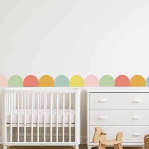 May include: A nursery scene featuring a white cot and chest of drawers. A colourful, semi-circular wall decal in pastel shades of pink, green, orange, yellow, and blue decorates the wall above the furniture. A wooden rocking horse sits in front of the drawers.