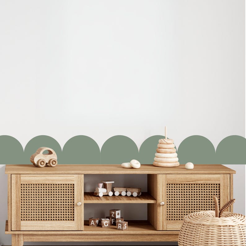 May include: A light wood cabinet with woven door fronts, displaying wooden toys. A wooden car, a train, and a stacking ring toy are visible. The cabinet is set against a white wall with a green scalloped border.