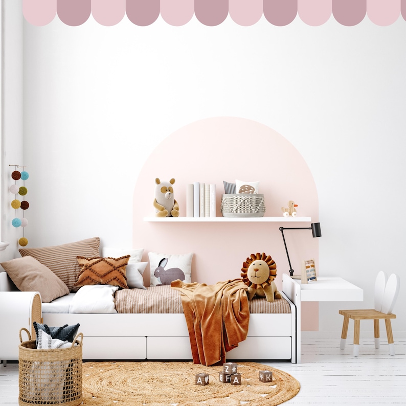 May include: A child's bedroom scene featuring a white bed with cushions and a brown blanket. A plush lion and bear are on the bed. A small desk and chair are to the right. A woven rug and basket are in the foreground. The room has a pink accent wall.