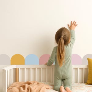 May include: A child in a sage green onesie reaches towards a wall decorated with pastel-coloured semi-circles. The child is standing near a white cot with a peach-coloured blanket and a yellow pillow.