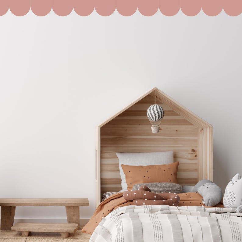 May include: A wooden house-shaped bed frame with a striped comforter, pillows, and soft toys. A small wooden bench is in front of the bed. A hot air balloon mobile hangs from the bed frame. The wall is white with a scalloped border.